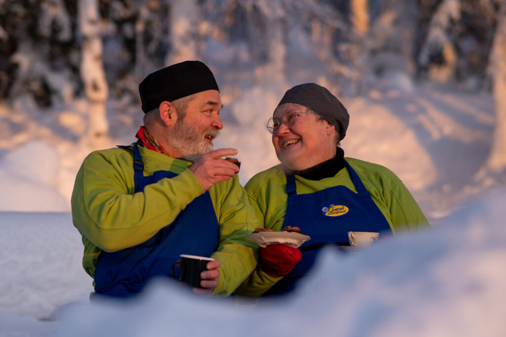 en leende man och en skrattande kvinna i grön tröja och blått förkläde sitter halvt dolda av en snöhög och dricker kaffe i solen.
