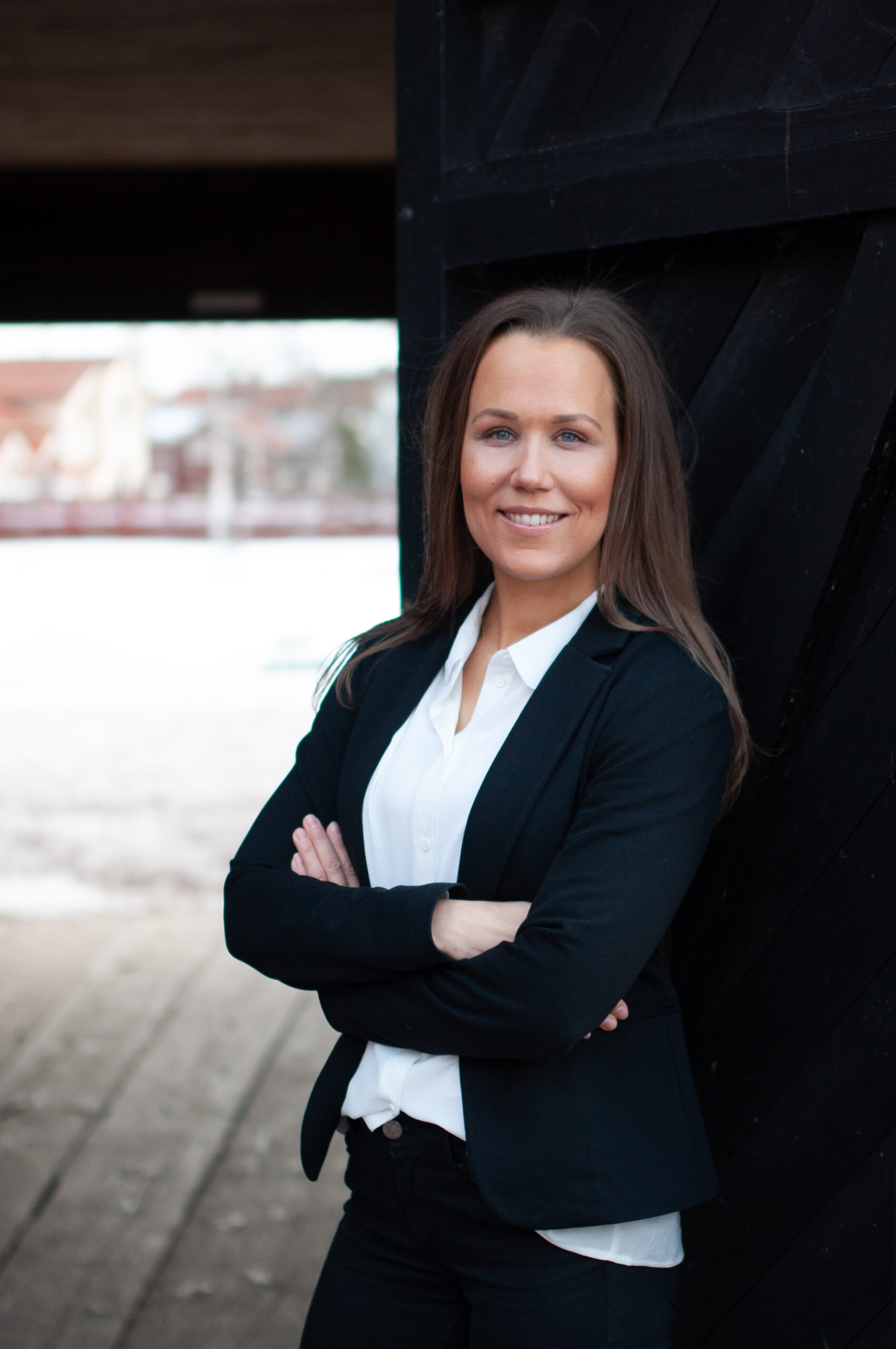 Kvinna med långt hår iklädd vit blus och kavaj står med armarna i kors och lutar sig mot en svart dörr