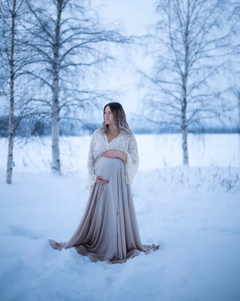 gravid kvinna i spets- och sidenklänning står i snöigt landskap under en gravidfotografering