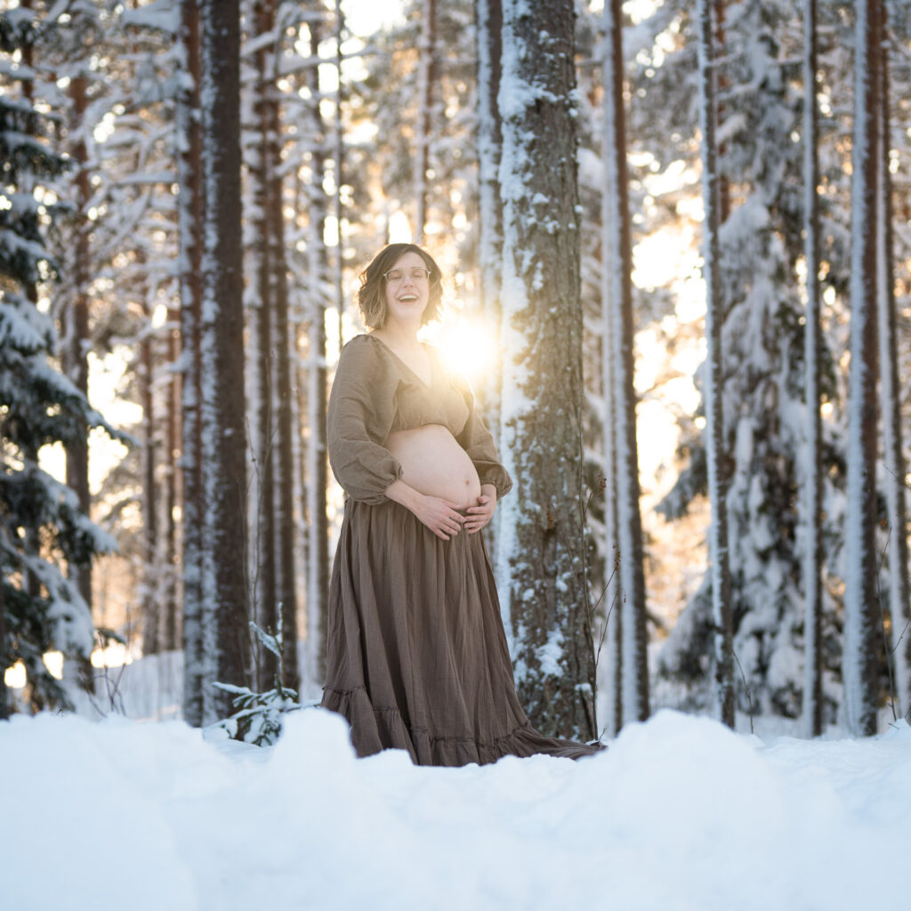 Gravid kvinna står i motljus på en snödriva. Hon är klädd i en brun tvådelad klänning så magen syns. Solen strålar bakom henne och ger henne ett glorialiknande sken