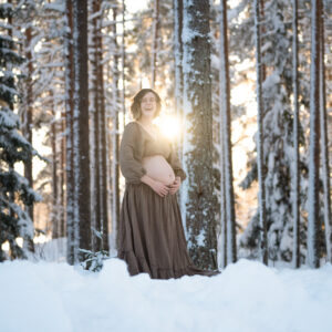 Gravid kvinna står i motljus på en snödriva. Hon är klädd i en brun tvådelad klänning så magen syns. Solen strålar bakom henne och ger henne ett glorialiknande sken