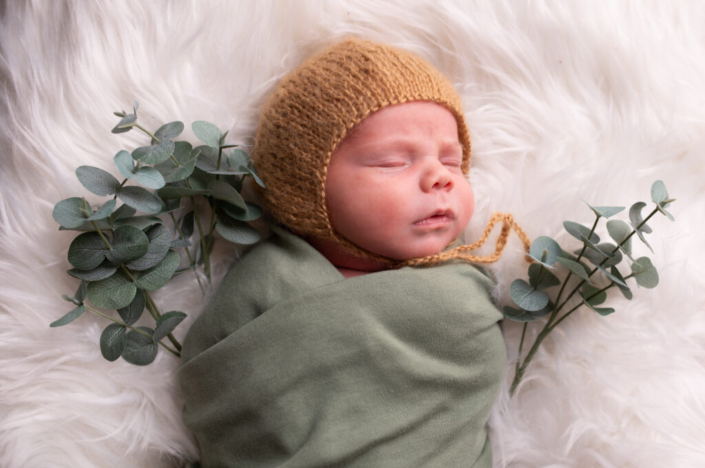 sovande nyfödd baby lindad i lindblomsgrön wrap. Bäbisen har en stickad hätta och vid sidan av ligger kvistar med gröna blad.