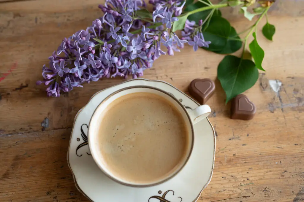 En lila syrenblomma och en kopp kaffe i gammalt porslin står på ett bord, brevid ligger två chokladhjärtan. Bilden är fotad som flatlay, uppifrån.