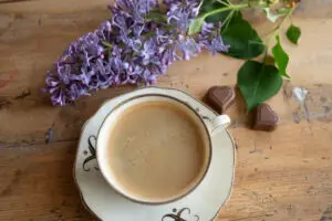 En lila syrenblomma och en kopp kaffe i gammalt porslin står på ett bord, brevid ligger två chokladhjärtan. Bilden är fotad som flatlay, uppifrån.