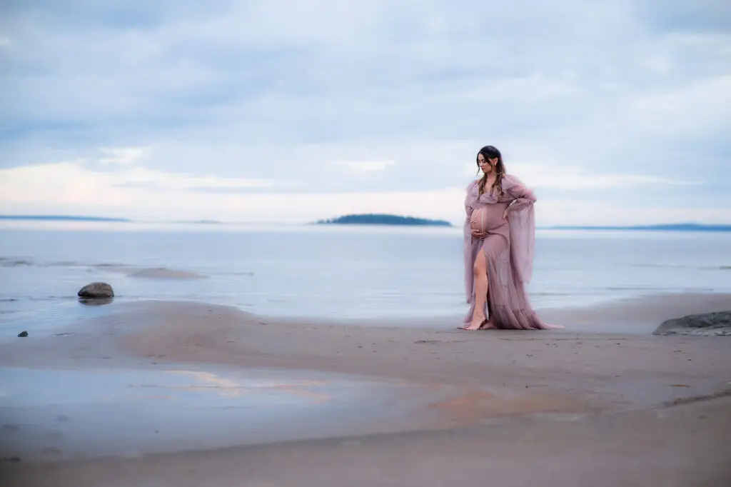 Gravid kvinna i lila tyllklänning står i vattenbrynet på en sandstrand och blickar ut över havet