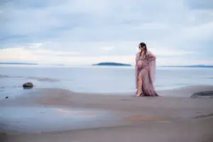 Gravid kvinna i lila tyllklänning står i vattenbrynet på en sandstrand och blickar ut över havet vid en gravidfotografering