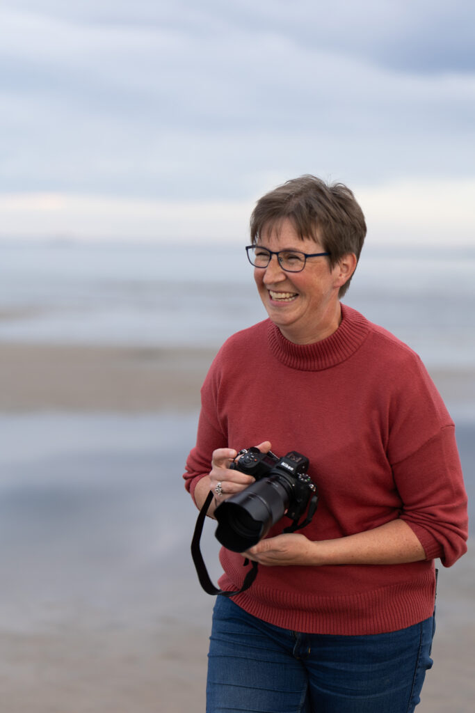 En glad fotograf i rödbrun tröja står på en strand med en kamera i handen