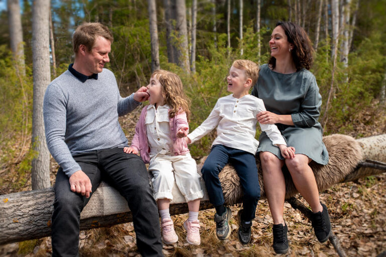 En skrattande familj med pappa, mamma, en pojke och en flicka sitter på en trädstam i skogen