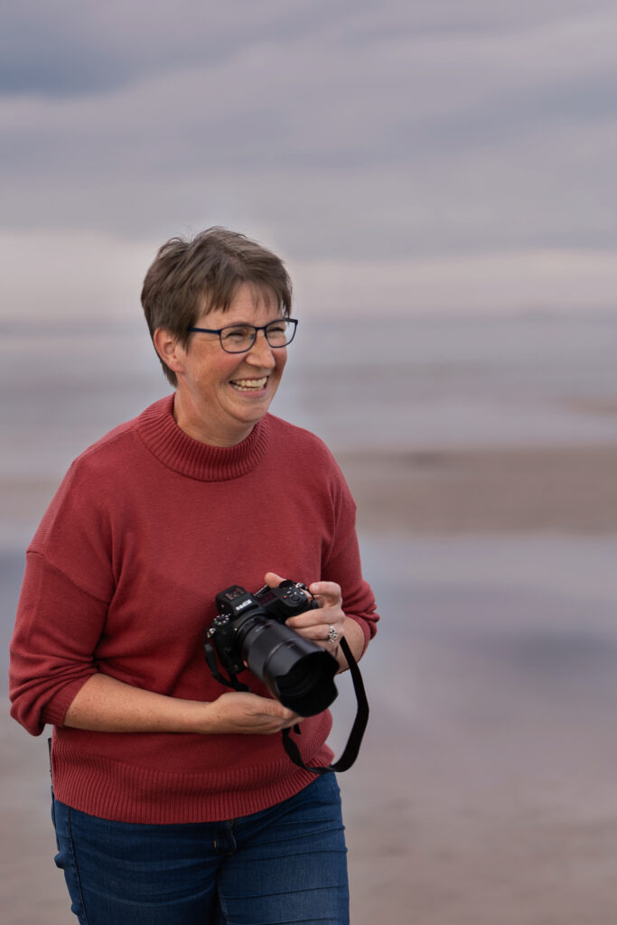En glad fotograf i rödbrun tröja står på en strand med en kamera i handen
