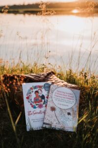 Produktbild av två böcker skrivna av Kicki Hansard, böckerna ligger mot en näverkont. I bakgrunden syns midnattssolen över an å