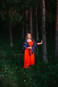 En kvinna i röd klänning och samisk sjal står i en tallskog och håller handen mot en trädstam