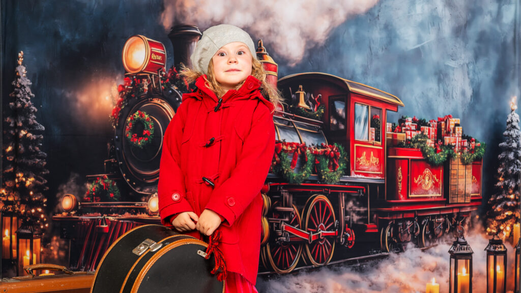 En lite flicka i röd kappa och svarta kängor står framför ett juligt ånglok. I händerna håller hon en hattast.