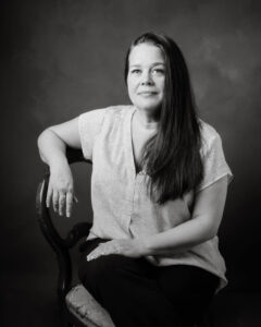 En kvinna med ljus kortärmad blus sitter och lutar ena armen mot ryggstödet på en stol. Kvinnan har långt mörkt hår som hänger fram över ena axeln, hon tittar avslappnat in i kameran.