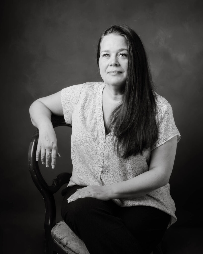 En kvinna med ljus kortärmad blus sitter och lutar ena armen mot ryggstödet på en stol. Kvinnan har långt mörkt hår som hänger fram över ena axeln, hon tittar avslappnat in i kameran.