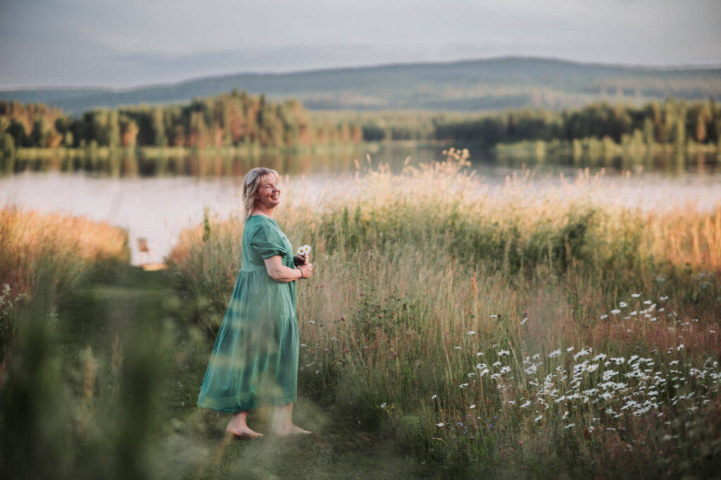 En leende kvinna i grön sommarklänning står på en stig på en äng med högt gräs