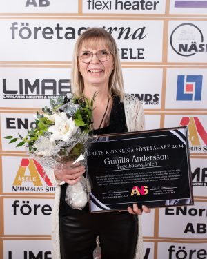 En glad kvinna med glasögon håller i en vinnarplakett och en blomsterbukett framför en sponsorbackdrop